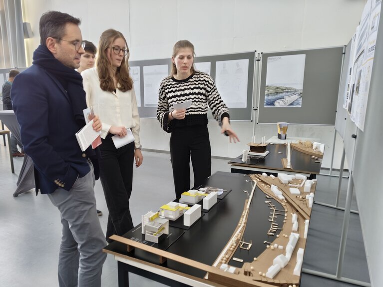 Ein Mann im Bild links und zwei Studentinnen stehen und betrachten Entwürfe der Architekturstudierendenn.