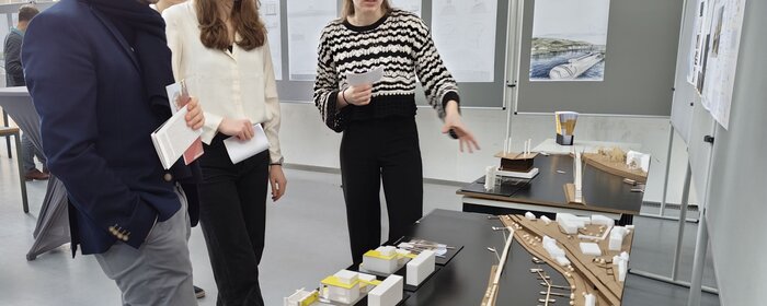 Ein Mann im Bild links und zwei Studentinnen stehen und betrachten Entwürfe der Architekturstudierendenn.