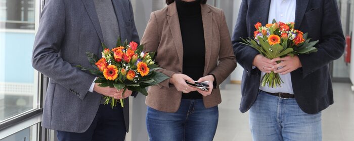 Zwei Männer stehen zusammen mit einer Frau in der Mitte. Die Männer halten jeweils einen Blumenstrauß in Ihrer Hand. 