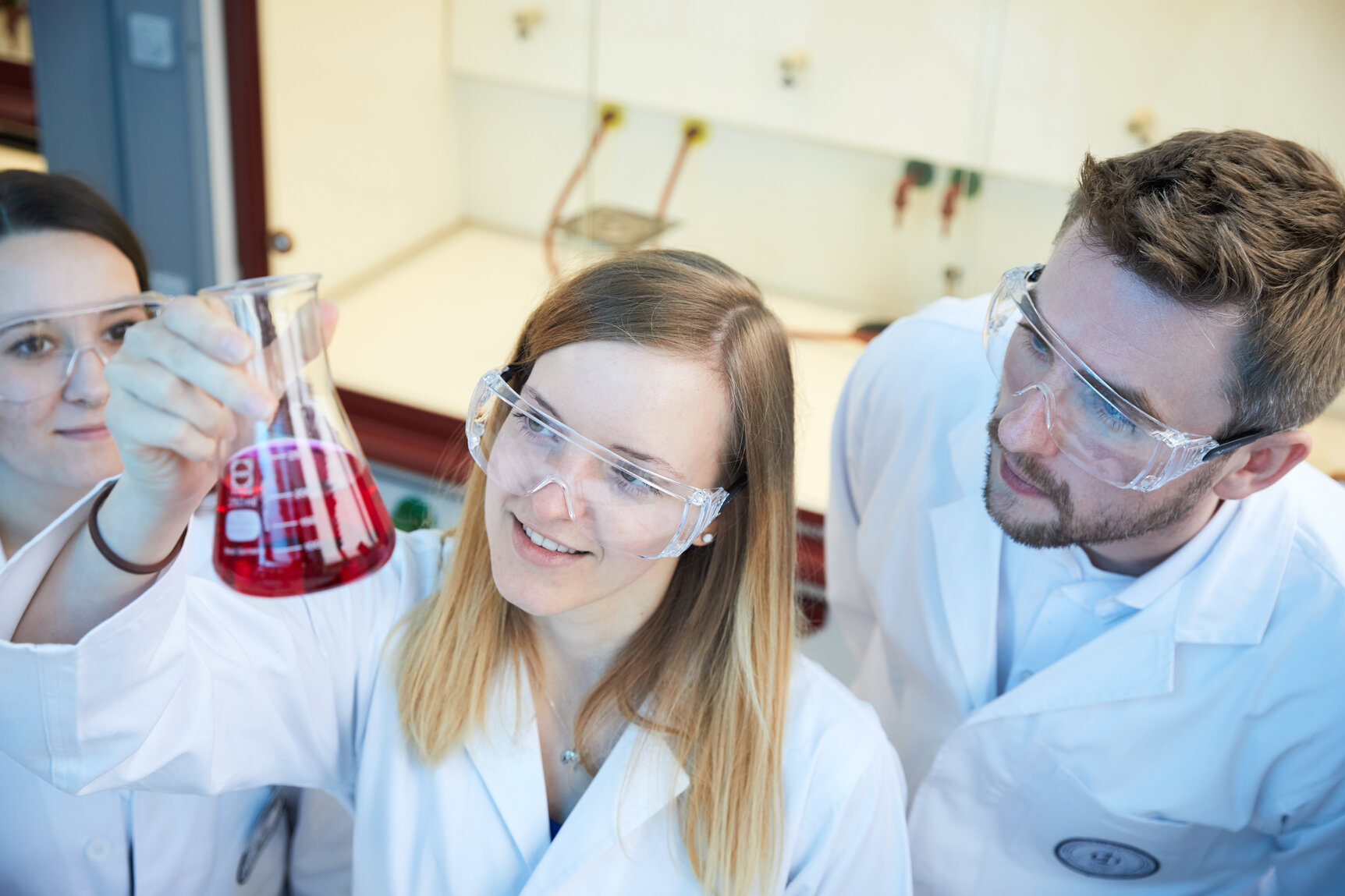 Dekoratives Bild, Studierende in weißen Kitteln und mit Schutzbrillen schauen auf ein Laborglas mit roter Flüssigkeit.
