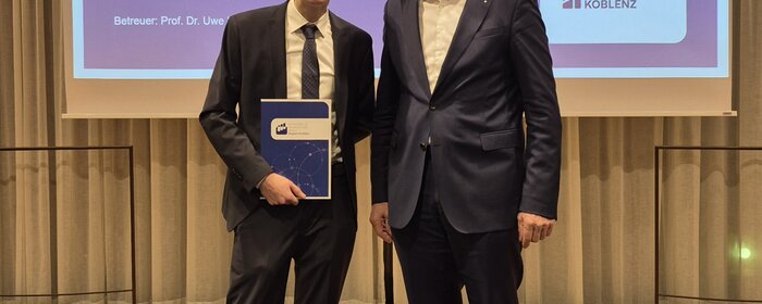 Zwei Männer, einer davon mit einer Urkunde in der Hand, vor einer Präsentationswand.