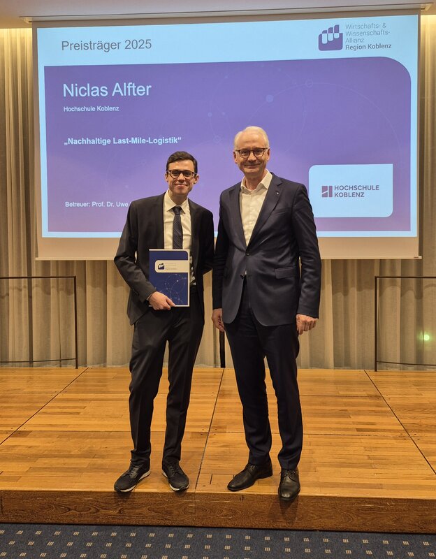 Zwei Männer, einer davon mit einer Urkunde in der Hand, vor einer Präsentationswand.