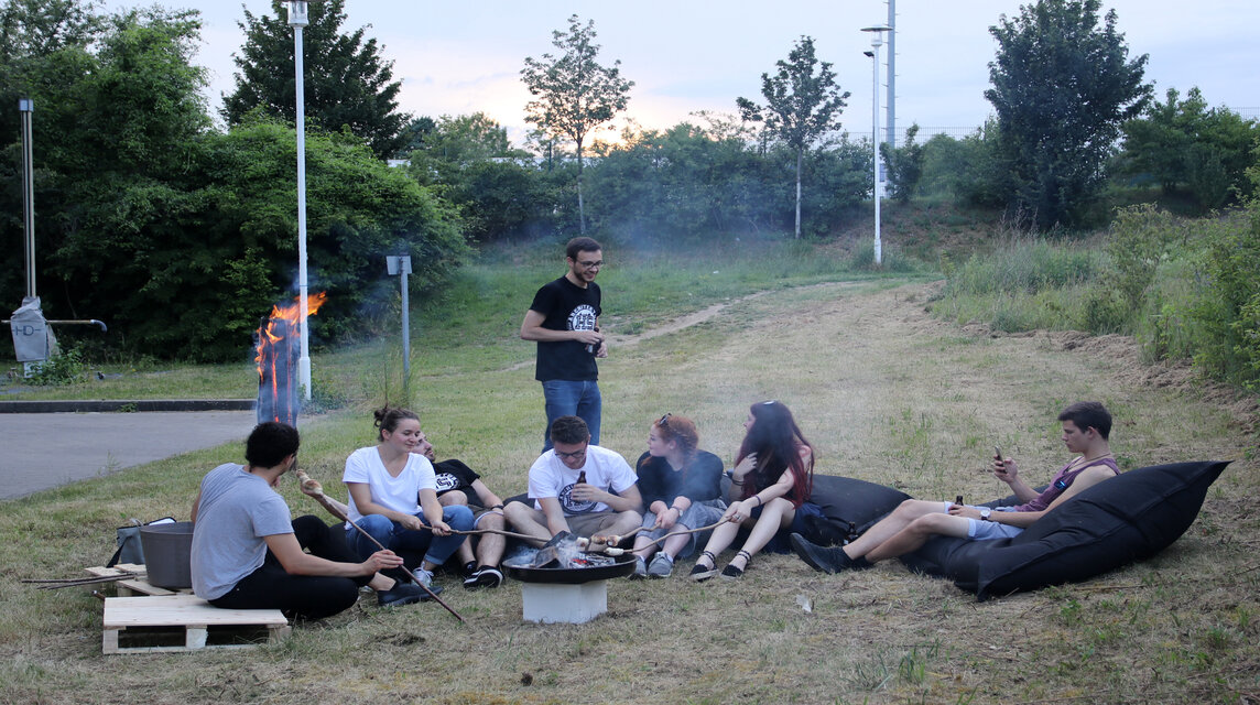 Studierende sitzen auf einer Wiese um einen Grill herum