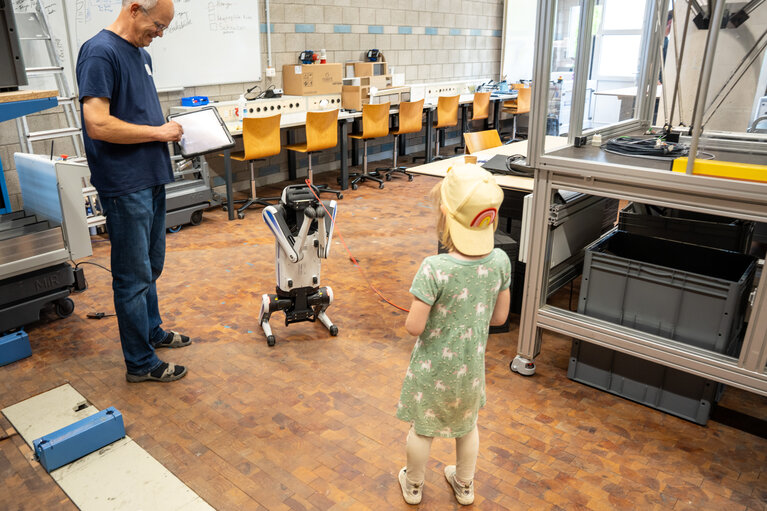 Mädchen mit Roboterhund an der Leine. Ein Mann mit Tablet daneben.