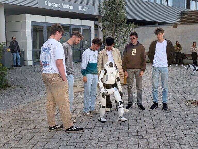 Humanoide Robotik Auf dem Vorplatz der Hochschule Koblenz stehen fünf Studierende gemeinsam mit einem humanoiden Roboter. Sie arbeiten an einem Projekt zur humanoiden Robotik und programmieren dabei den Roboter DOBOT LUMO L1. Der Roboter verfügt über mehr als 30 Freiheitsgrade und kann unter anderem seine Arme, Beine, Taille und seinen Kopf bewegen. Im Rahmen des Projekts lernen die Studierenden Grundlagen der Steuerung, Regelung und Programmierung und erproben, wie weit der Roboter selbstständig joggen kann.