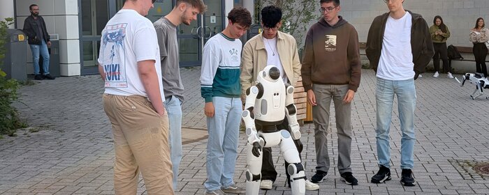 Auf dem Vorplatz der Hochschule Koblenz stehen fünf Studierende gemeinsam mit einem humanoiden Roboter. Sie arbeiten an einem Projekt zur humanoiden Robotik und programmieren dabei den Roboter DOBOT LUMO L1. Der Roboter verfügt über mehr als 30 Freiheitsgrade und kann unter anderem seine Arme, Beine, Taille und seinen Kopf bewegen. Im Rahmen des Projekts lernen die Studierenden Grundlagen der Steuerung, Regelung und Programmierung und erproben, wie weit der Roboter selbstständig joggen kann.