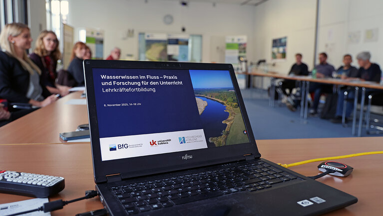 Ein Laptop steht auf einem Tisch. Unscharf sind im Hintergrund Menschen in einem Sitzungssaal zu sehen.