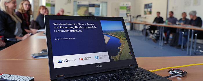 Ein Laptop steht auf einem Tisch. Unscharf sind im Hintergrund Menschen in einem Sitzungssaal zu sehen.