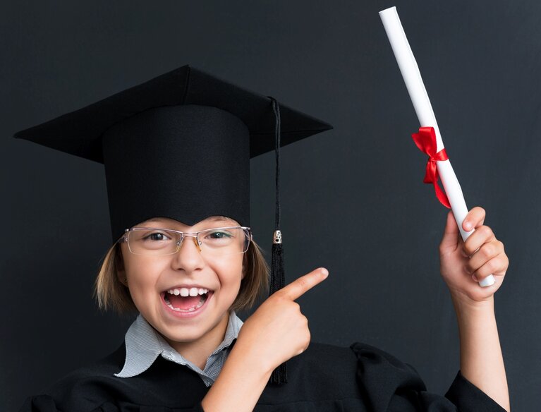 Ein Kind mit Doktorhut und einem gerollten weißen Blatt mit rotem Band in der Hand.