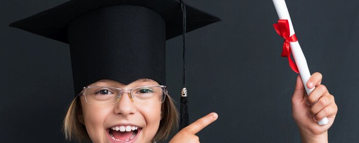 Ein Kind mit Doktorhut und einem gerollten weißen Blatt mit rotem Band in der Hand.