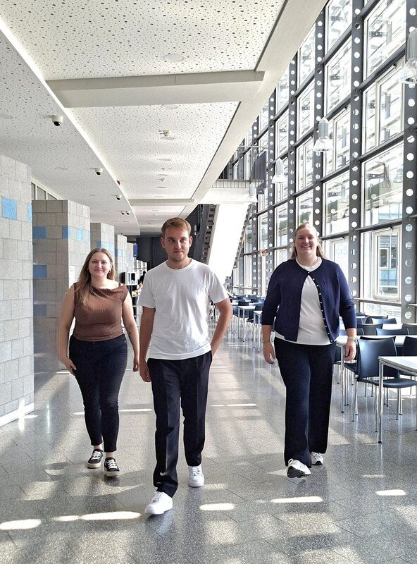 Zwei Frauen und ein Mann (Mitte) gehen einen Gang an der Hochschule entlang und laufen auf die Kamera zu.