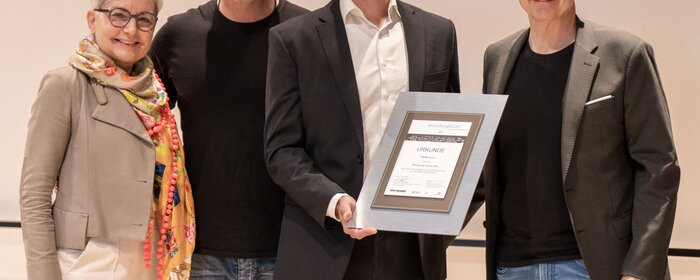 Vier Personen stehen zusammen. Der Preisträger hält eine Urkunde.