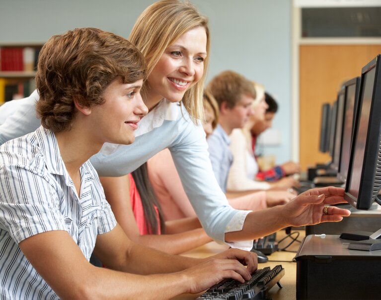 Ein junger Mann am Computer. Eine Frau steht, zeigt auf den Bildschirm und erklärt.