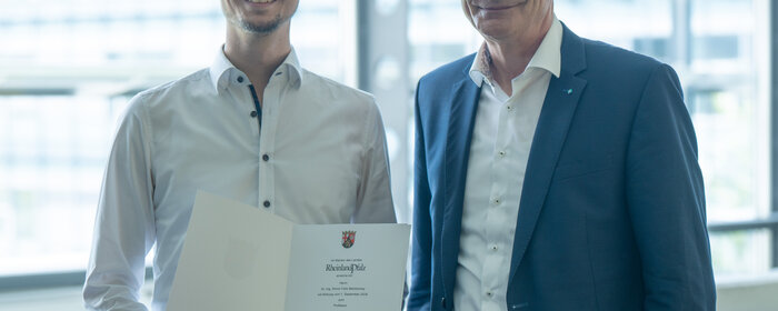 Zwei Herren stehen beieinander. Der Linke von Beiden hält eine Urkunde in der Hand.