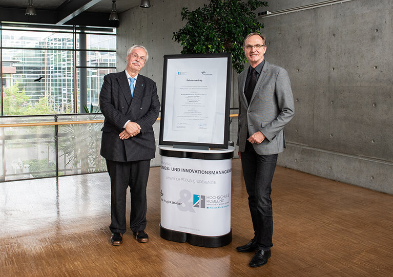 Vertrag zwischen dem DLR Projektträger und der Hochschule Koblenz steht im Rahmen auf einem Tisch. Links daneben steht Prof. Dr. Kristian Bosselmann-Cyran und rechts daneben Klaus Uckel, Gschäftsleiter DLR-Projektträger