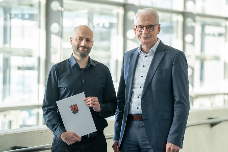 Zwei Männer stehen zusammen. Der linke von ihnen hält eine Urkunde.