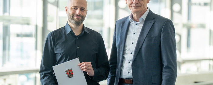 Zwei Männer stehen zusammen. Der linke von ihnen hält eine Urkunde.