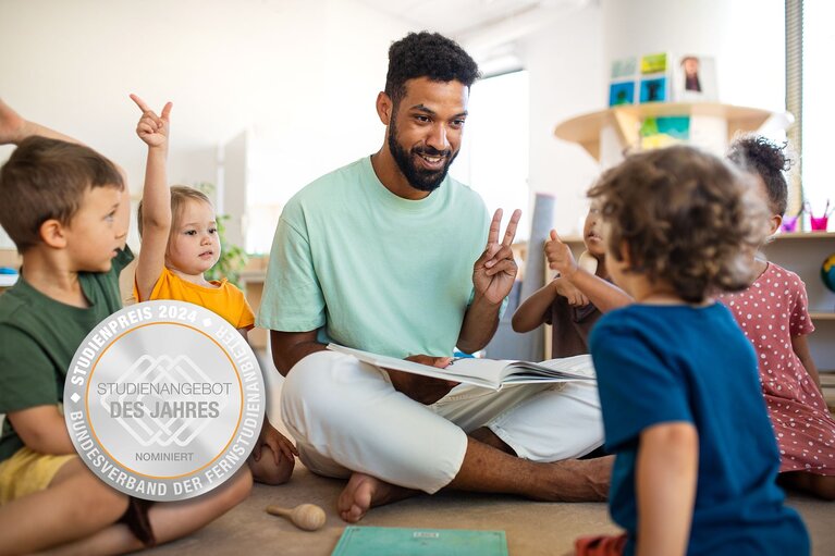 Lachender Erzieher sitzt mit einem Buch inmitten einer Kindergruppe auf dem Boden und hält zwei Finger hoch