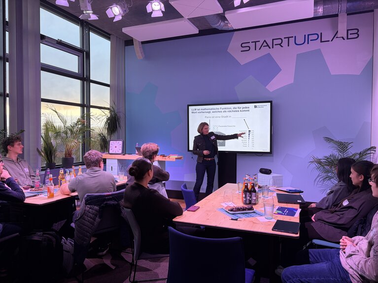 Eine Frau steht im Seminarraum vorne und erklärt etwas an der Präsentationswand. Jugendliche sitzen an Tischen.