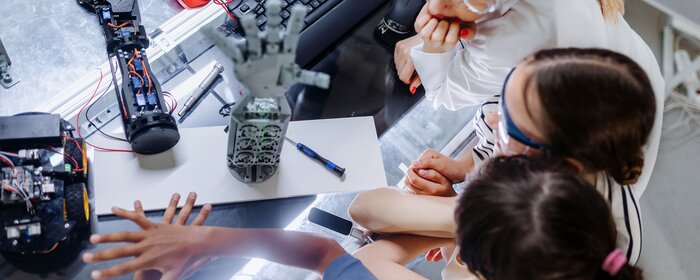 Drei Mädchen und eine Frau sitzen an einem Experimentiertisch. Darauf stehen Roboterhände.