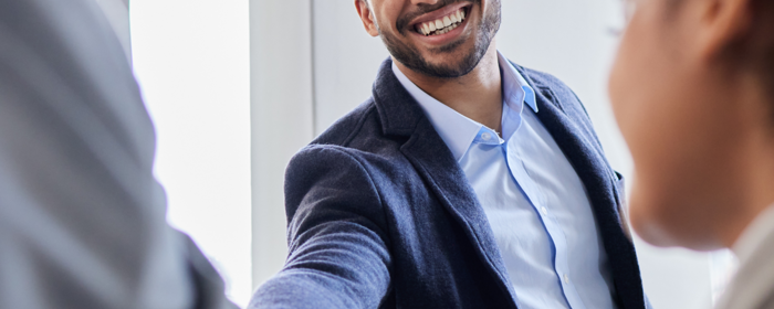 Ein Mann gibt einer anderen Person, von der man nur den Arm sieht, die Hand. Eine andere Person ist seitlich im Bild.