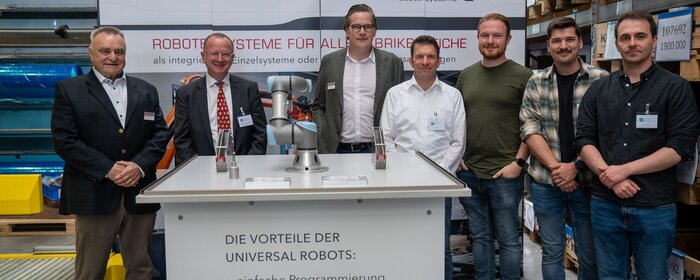 Sieben Männer und ein Roboter vor einem Präsentationstisch