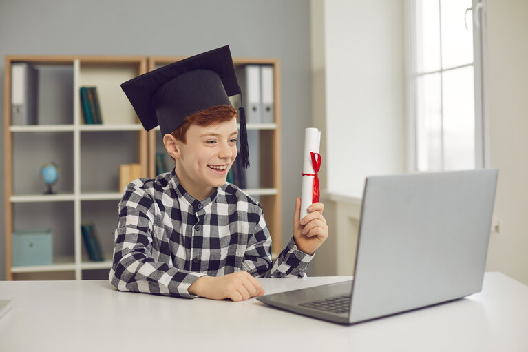Graduate boy sitting at desk in graduate hat and holding scroll of diploma in front of laptop.
