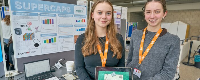 Zwei Mädchen stehen zusammen an ihrem Projektstand und halten ein iPad mit einer Titelfolie ihres Projekts hoch. 