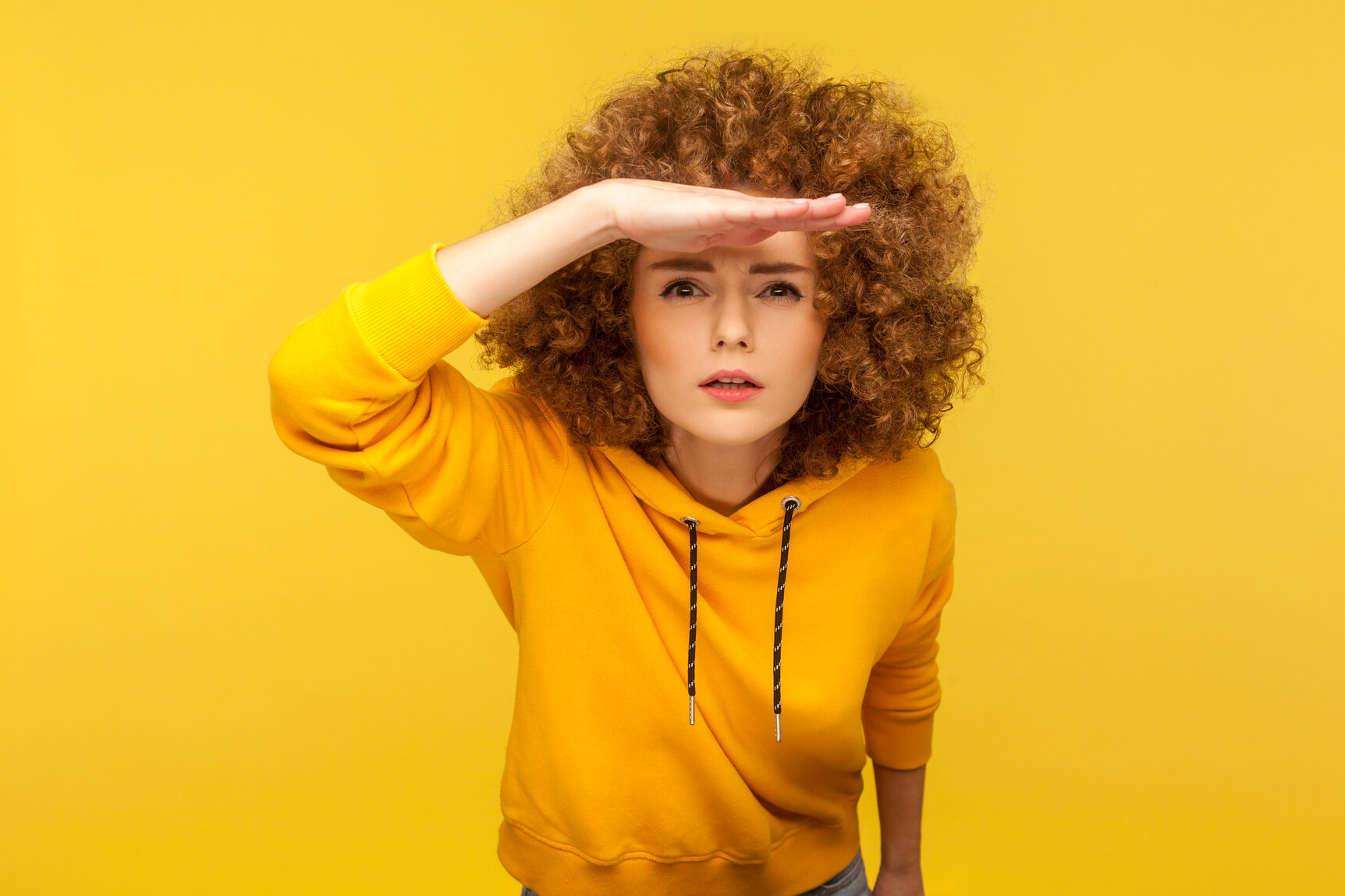 Portrait of curious woman in urban style hoodie holding hand above eyes and peering into distance