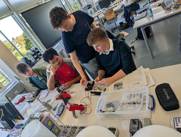 Experimentieren und Messen Drei Studierende arbeiten gemeinsam an einem elektrotechnischen Experiment. An einem Tisch mit Experimentierboxen, elektronischen Bauteilen und Messgeräten führen sie unter Anleitung einer wissenschaftlichen Hilfskraft praktische Messungen durch. Die wissenschaftliche Hilfskraft erklärt den Studierenden die Vorgehensweise und unterstützt sie bei der Durchführung des Experiments. Die Studierenden betrachten aufmerksam die Versuchsanordnung und arbeiten gemeinsam an der Lösung der Aufgabe.