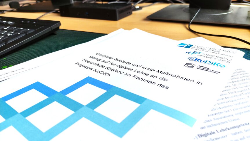 Das Bild zeigt einen Stapel Papier auf einem Schreibtisch. Auf dem obersten Blatt ist der Titel „Ermittelte Bedarfe und erste Maßnahmen in Bezug auf die digitale Lehre an der Hochschule Koblenz im Rahmen des Projektes KuDiKo“ zu erkennen.