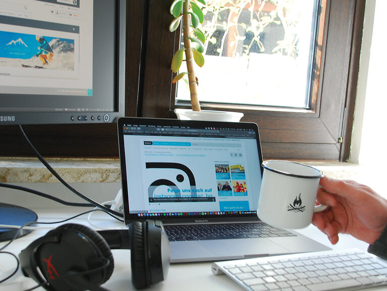 Schreibtisch mit Bildschirm, Laptop und Kopfhörern. Im Vordergrund eine Hand mit einem Kaffeebecher. So sehen Home Office Arbeitsplätze für Studierende aus