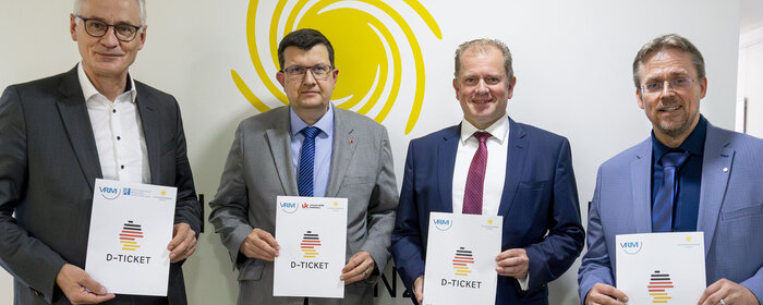 Vier Männer stehen vor einer weißen Wand mit dem gelben Logo des Studierendenwerks und halten den Vertrag zum Deutschland-Semesterticket in der Hand