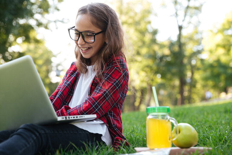 Happy brunette schoolgirl in eyeglasses sitting on grass with juice