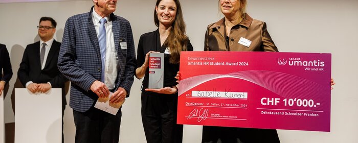 Zwei Frauen und ein Mann auf der Bühne. Die eine Frau hält einen Preis, die andere den Gewinnercheck. 