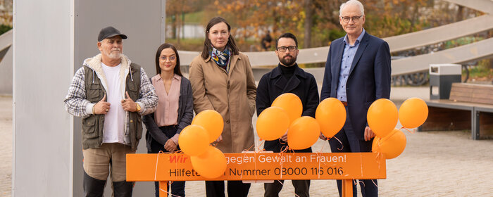 Fünf Personen stehen hinter einer orangen Bank. Daran sind orange Luftballons befestigt und auf der Bank ein Aufdruck mit der Notruf-Telefonnummer 08000116016 für Gewalt gegen Frauen.