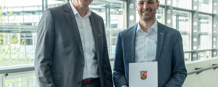 Zwei Herren stehen zusammen. Der rechte von beiden hält eine Urkunde in der Hand.
