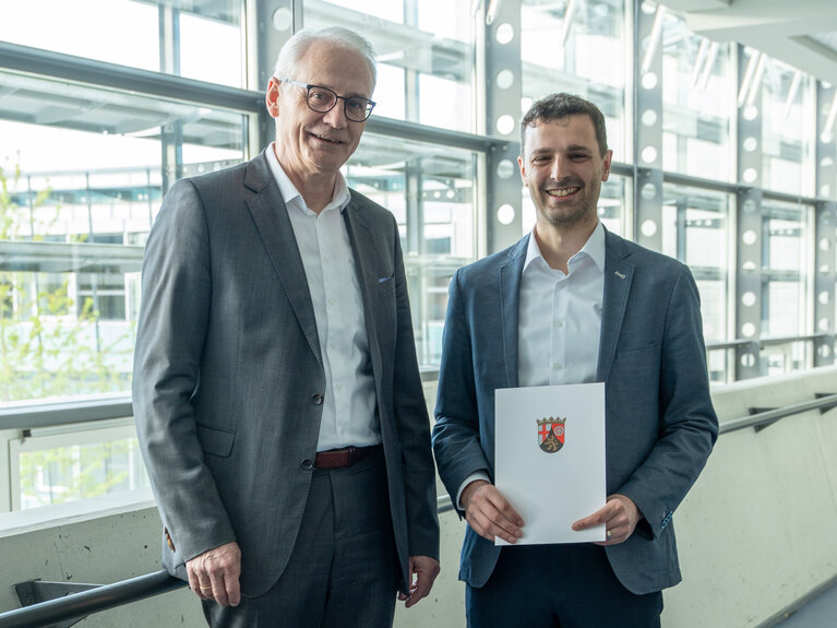 Zwei Herren stehen zusammen. Der rechte von beiden hält eine Urkunde in der Hand.