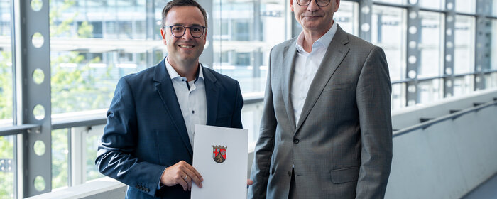 Zwei Männer stehen zusammen. Der linke von beiden hält eine Urkunde in der Hand.