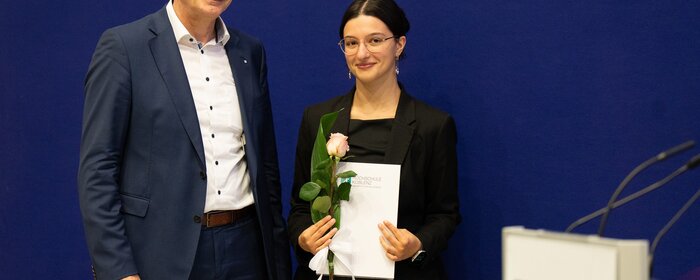 Ein Herr und eine Dame stehen vor dem Banner der Hochschule Koblenz mit Auszeichnung und Blumen in der Hand.