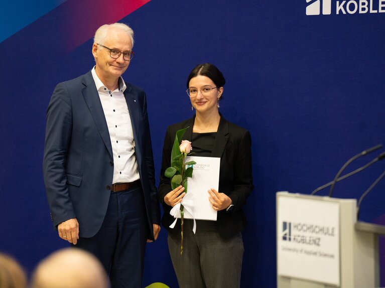 Ein Herr und eine Dame stehen vor dem Banner der Hochschule Koblenz mit Auszeichnung und Blumen in der Hand.