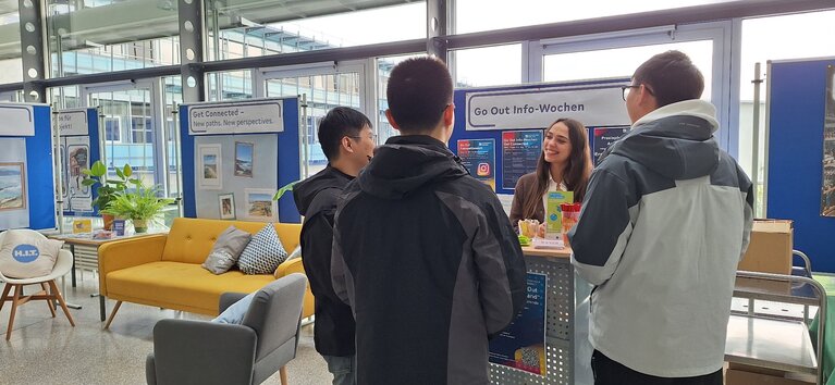 Drei Studierende stehen bei einer Beraterin des International Office am Infostand.