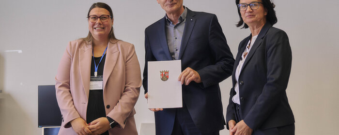Zwei Frauen und ein Mann (Mitte) stehen zusammen. Der Mann hält eine Urkunde.