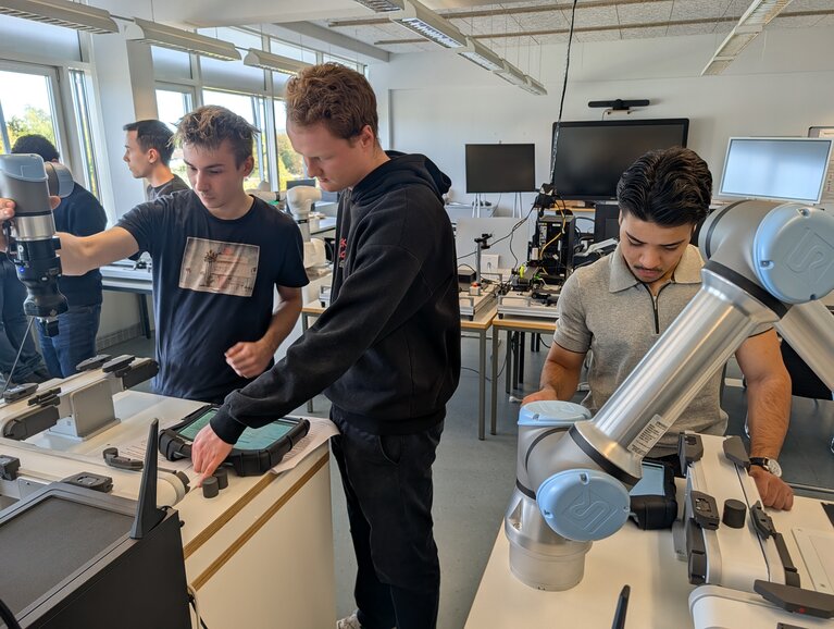Roboterprogrammierung am UR5e Die Erstsemesterstudierenden - hier fünf an der Zahl - arbeiten gemeinsam an einem kollaborativen sechsachsigen UR5e-Roboter. Sie lernen dabei die Grundlagen der Roboterprogrammierung und üben unter anderem die Bewegung des Roboters im kartesischen Raum sowie die Bearbeitung von Werkstücken und die Ansteuerung von Ein- und Ausgängen.