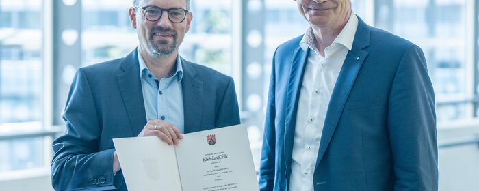 Zwei Männer stehen beieinander. Ein Mann hält eine Urkunde.