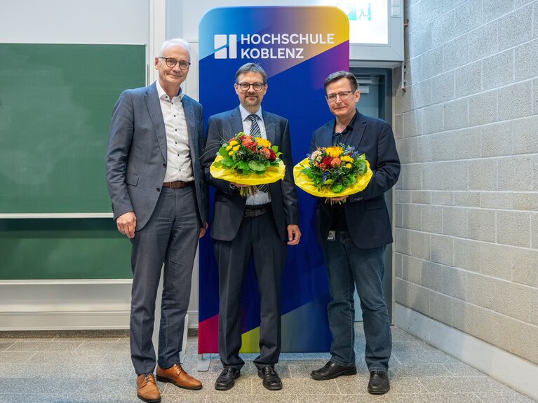 Drei Männer stehen zusammen vor einem Aufsteller mit dem Schriftzug Hochschule Koblenz. Die beiden rechten Herren halten einen Blumenstrauß in ihren Händen.