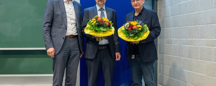 Drei Männer stehen zusammen vor einem Aufsteller mit dem Schriftzug Hochschule Koblenz. Die beiden rechten Herren halten einen Blumenstrauß in ihren Händen.