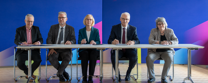 Drei Männer und zwei Frauen sitzen an einem Tisch zur Vertragsunterzeichnung. Im Hintergrund Stellwände der Hochschule Koblenz