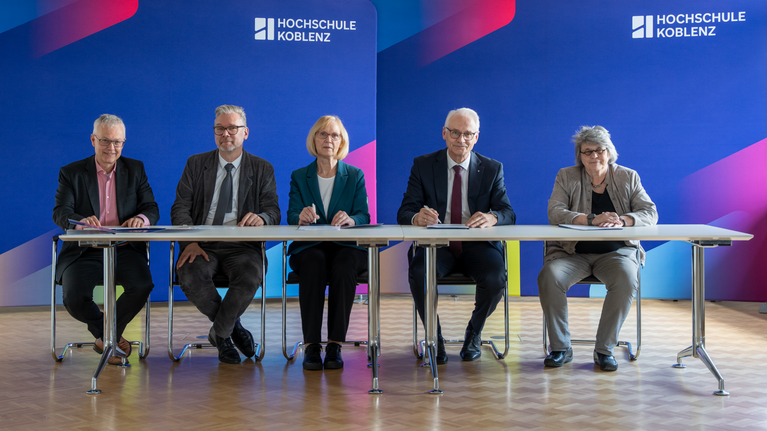 Drei Männer und zwei Frauen sitzen an einem Tisch zur Vertragsunterzeichnung. Im Hintergrund Stellwände der Hochschule Koblenz