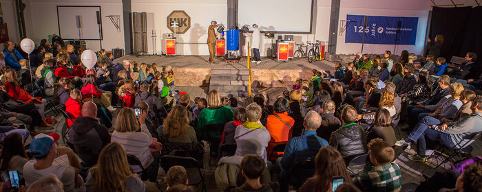 Eine teilnehmende Großgruppe von hinten mit Blick auf die Bühne, wo im Rahmen der Nacht der Technik Experimente vorgeführt werden.